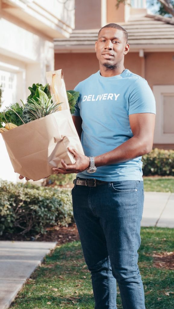 Smiling delivery man holding a paper bag of groceries on a sunny day outdoors.